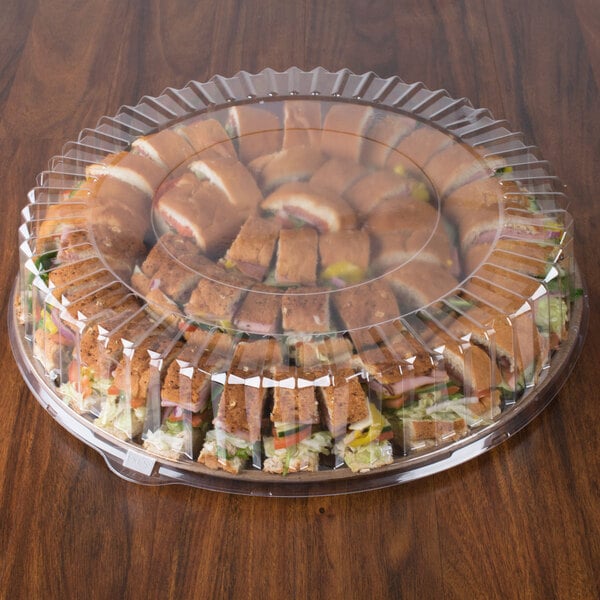 A clear Solut round plastic catering tray lid over a table of sandwiches.