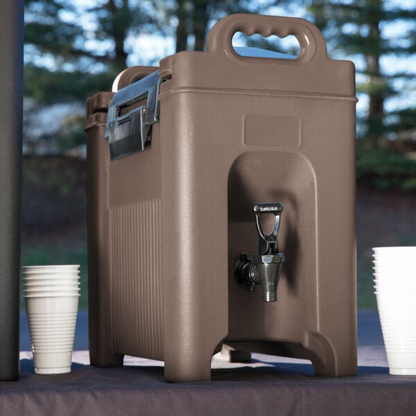 A brown plastic Carlisle Cateraide XT insulated beverage dispenser with a faucet and white cups.