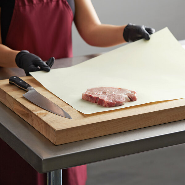 A person wearing black gloves using a knife to cut meat on a white Gardenia Premium paper-covered cutting board.