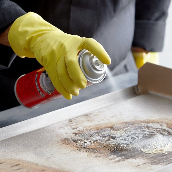 A person in yellow gloves using a CARBON-OFF spray can to clean a pan in a professional kitchen.
