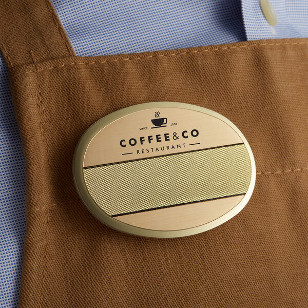 A brown apron with a customizable gold oval metal nametag on the counter in a coffee shop.