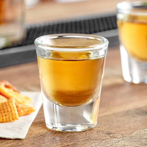 An Acopa shot glass filled with brown liquid next to a napkin with crackers and a snack.