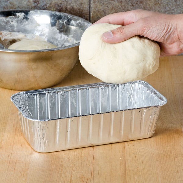 A hand holding a ball of dough over a Durable Packaging foil bread loaf pan.