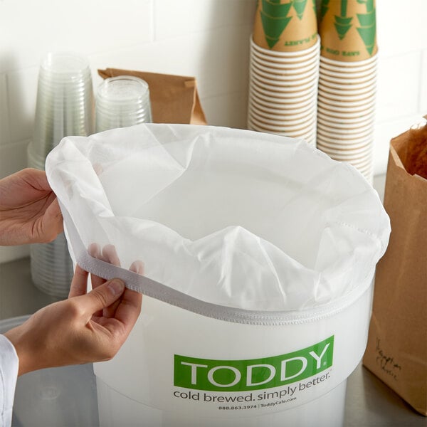 A hand holding a Toddy commercial cold brew strainer over a container with a white plastic bag inside.