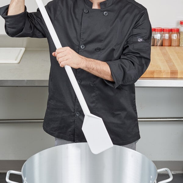 A man in a chef's uniform using a Carlisle white nylon paddle with a polypropylene handle to stir a large pot.