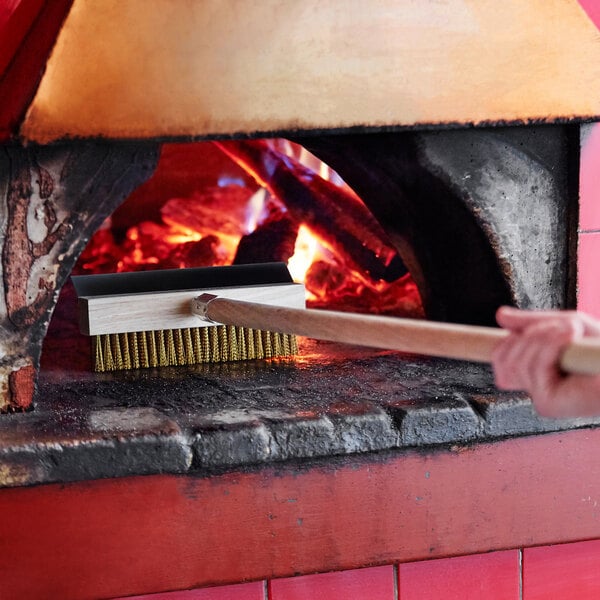 A person using an American Metalcraft pizza oven brush with a scraper to clean a pizza oven.