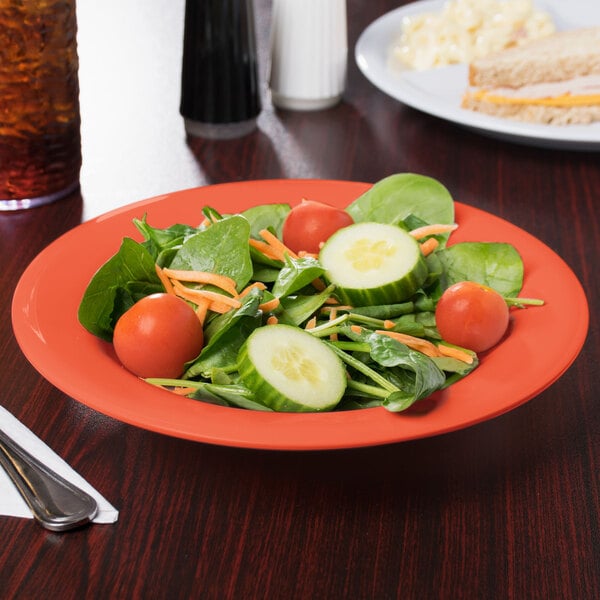 A bowl of salad with tomatoes, cucumbers, and carrots on a table with a sandwich.