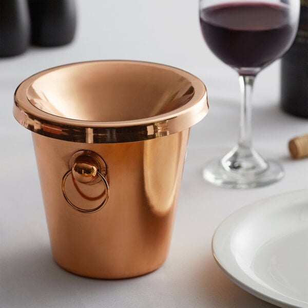 A copper Franmara wine tasting spittoon with a handle next to a glass of wine.