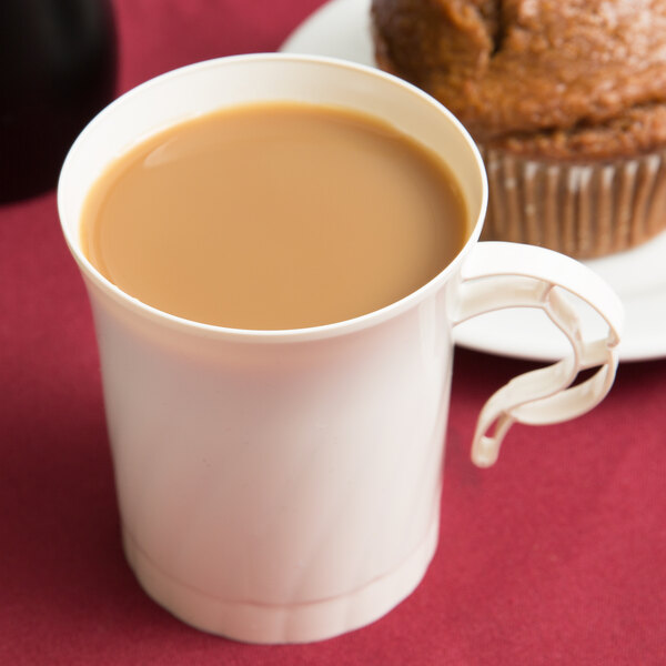 An ivory WNA Comet disposable plastic coffee cup filled with coffee next to a muffin on a white plate.