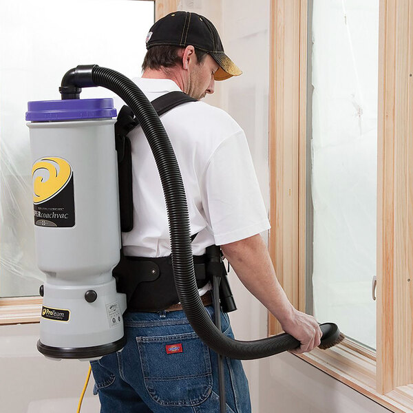 A man in a white shirt and blue jeans uses a ProTeam Super CoachVac backpack vacuum to clean a room.