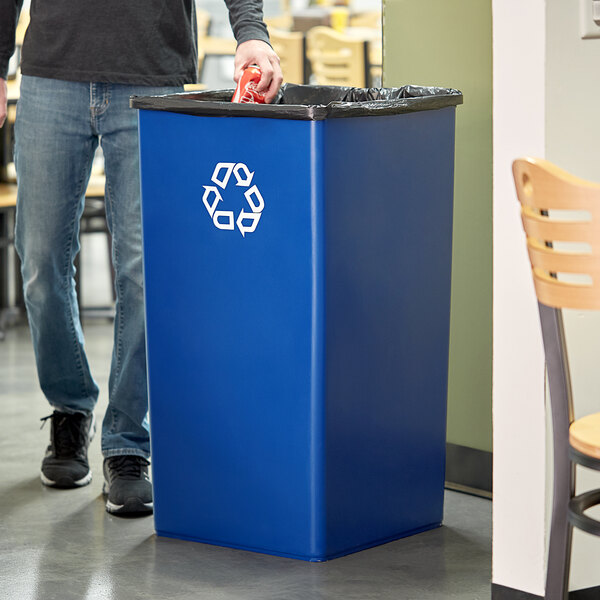 A man standing next to a Rubbermaid blue square recycling bin with a recycle symbol on it.