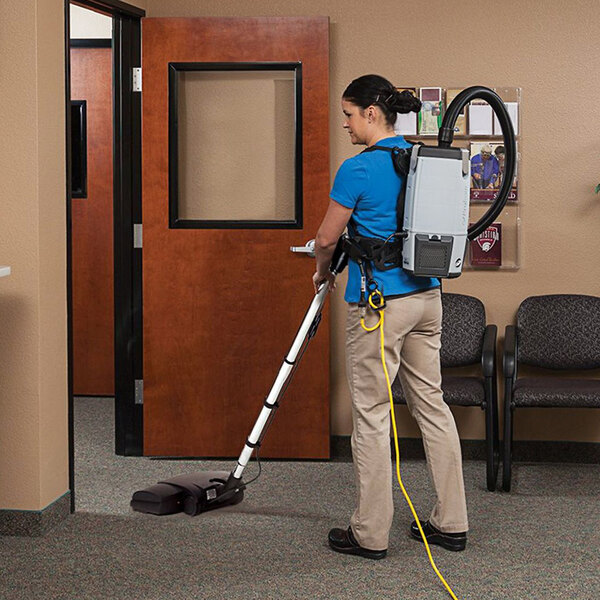 A woman using a ProTeam backpack vacuum with black and white accents to clean an office cafeteria.