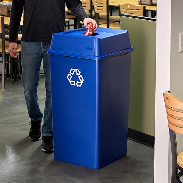 A man standing next to a blue Rubbermaid recycle bin with a bottle/can hole lid.
