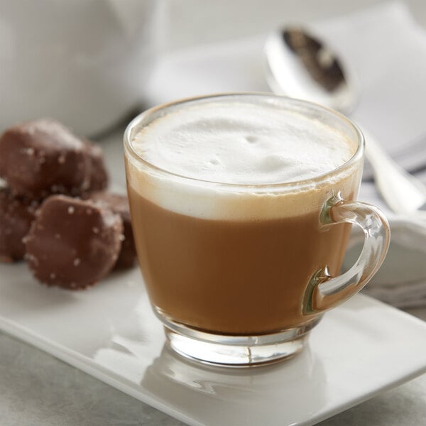A glass cup of UPOURIA hot chocolate with white foam and chocolate candies on top.