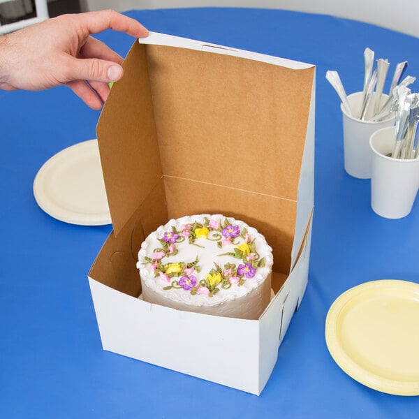 A hand holding a 8" x 8" white cake box with a cake inside.