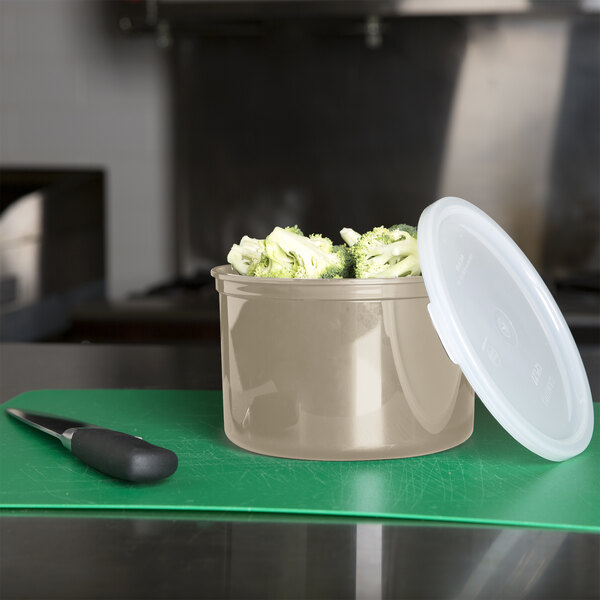 A beige Cambro round plastic crock with lid on a cutting board with broccoli and a knife.