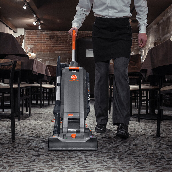 A man pushing a Hoover HushTone commercial vacuum cleaner in a restaurant dining area.
