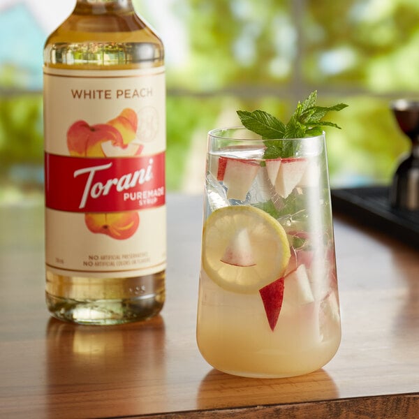 A glass of fruity drink with fruit slices next to a bottle of Torani White Peach syrup.