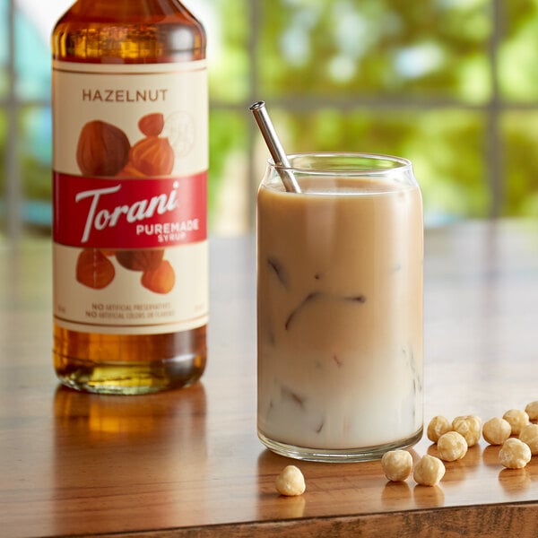 A glass of brown liquid with a metal straw next to nuts flavored with Torani Puremade Hazelnut Syrup.