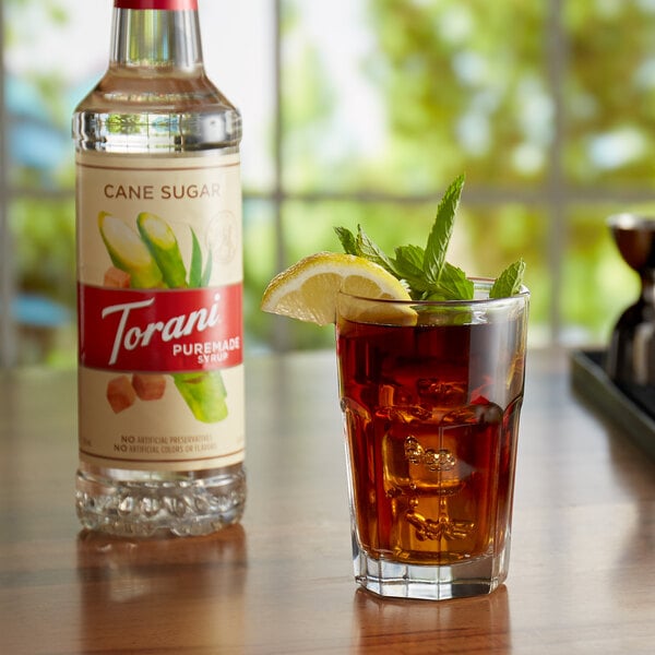 A glass of iced tea with mint leaves and a lemon wedge next to a Torani Pure Cane Sugar syrup bottle.