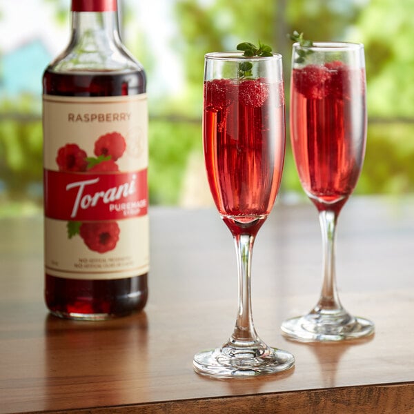 Two glasses of red liquid and a bottle of Torani Puremade Raspberry Flavoring Syrup.