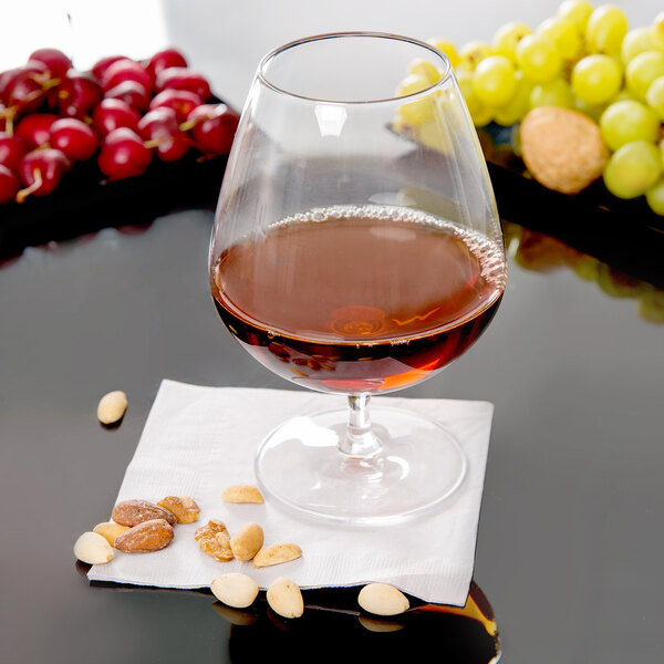 A Stolzle brandy snifter filled with brown liquid on a table next to grapes and nuts.