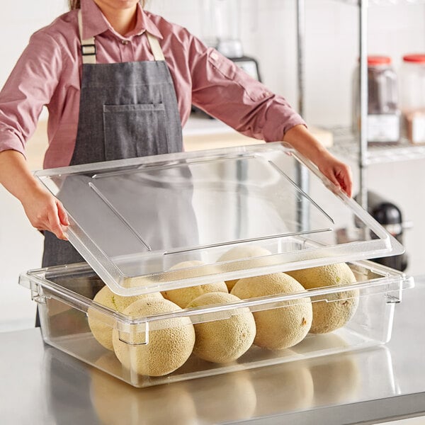 A woman in an apron holding a clear Cambro food storage container with a clear lid full of food.