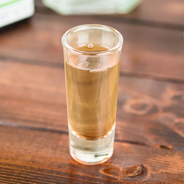 A Libbey tequila shooter glass filled with brown liquid on a wooden surface.