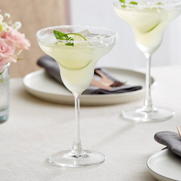 Two Acopa Covella margarita glasses filled with yellow liquid and garnished with lime slices on a table.