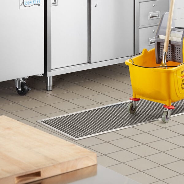 A yellow mop and bucket in a school kitchen with a stainless steel floor trough.