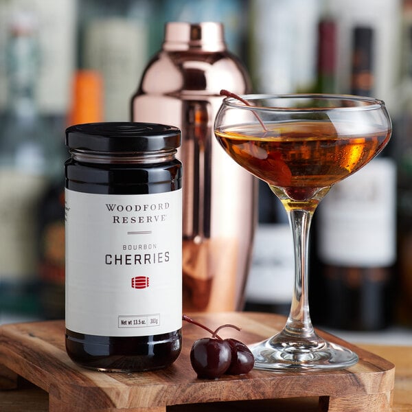A jar of Woodford Reserve Bourbon Cherries on a wooden tray with a glass of liquid.