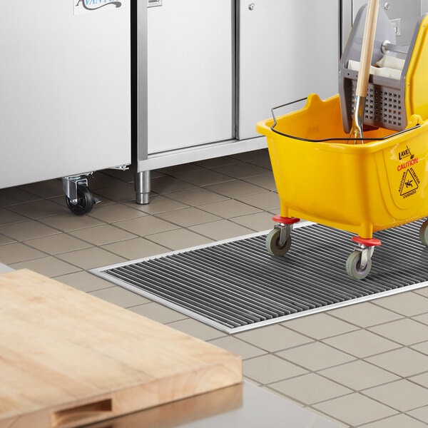 A yellow mop bucket sits on a Regency stainless steel floor trough in a kitchen.