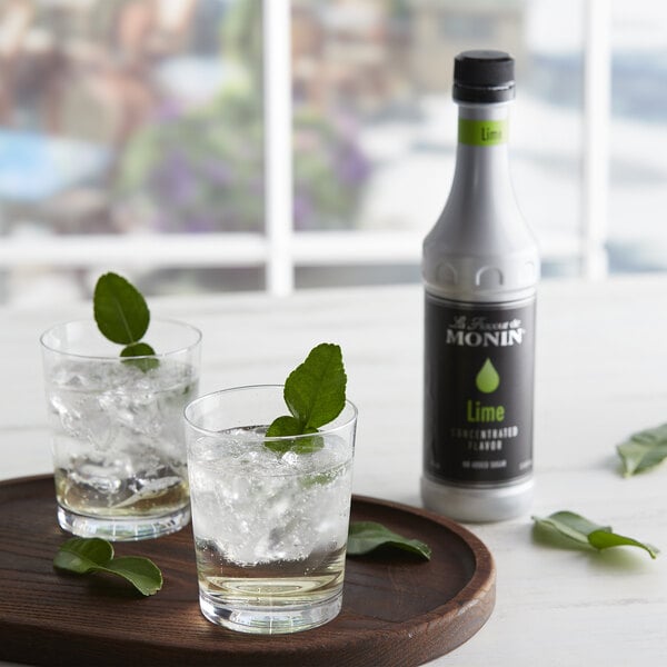 A bottle of Monin Lime Concentrated Flavor with two glasses of ice and a leafy drink.