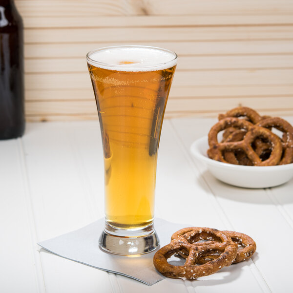 A Libbey pilsner glass of beer on a table with a bowl of pretzels.