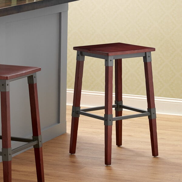 Two Lancaster Table & Seating rustic industrial bar stools with mahogany finish wooden legs next to a counter.