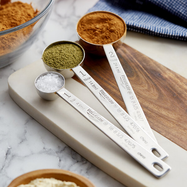 A set of Vollrath stainless steel measuring spoons with different spices on a counter.
