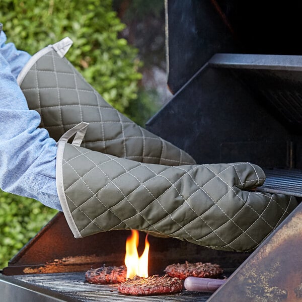 A person cooking meat on a grill using green Choice flame-retardant oven mitts.