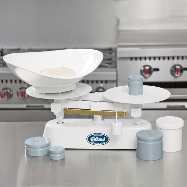 A white Edlund baker's dough scale on a counter with measuring cups and containers.