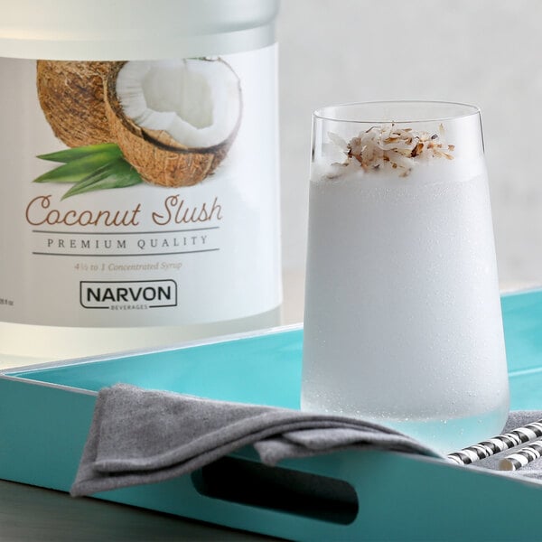 A white container of Narvon Coconut Slushy concentrate on a tray with a glass of coconut slush and straws.