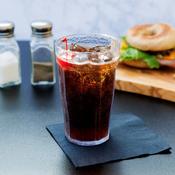 A Cambro clear plastic tumbler filled with soda, ice, and a cherry on a table with a bagel sandwich.