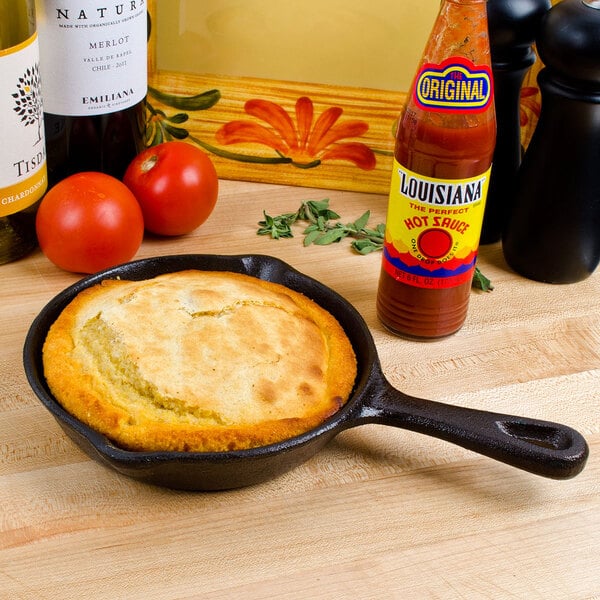 An American Metalcraft pre-seasoned mini cast iron skillet with cornbread next to a bottle of hot sauce.