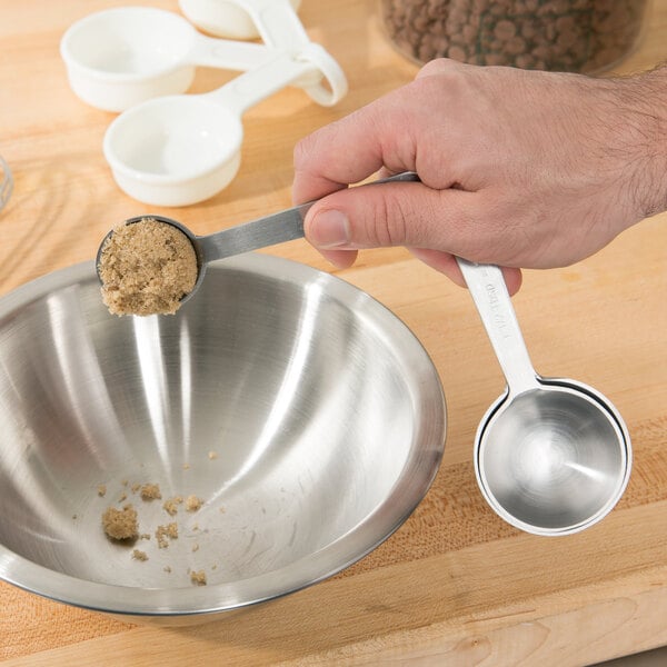 A hand holding Tablecraft stainless steel measuring spoons over a white bowl with brown sugar in one.