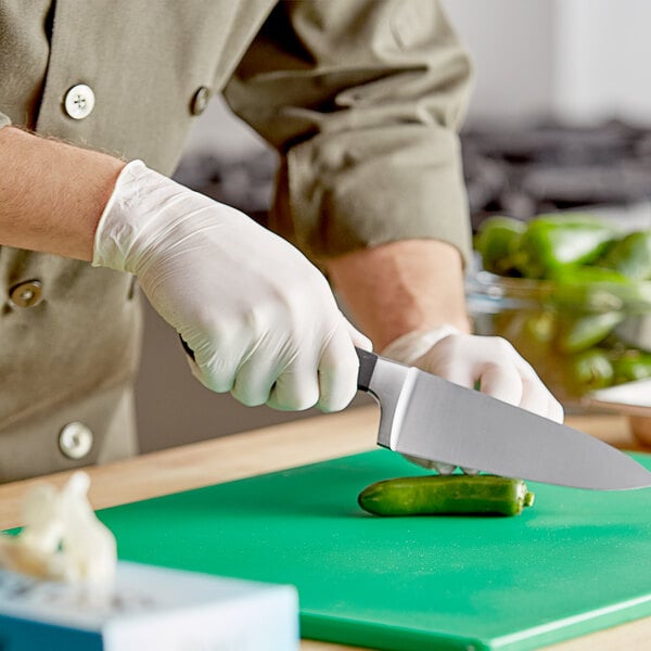A person wearing disposable gloves using a knife to cut a cucumber on a counter in a professional kitchen.