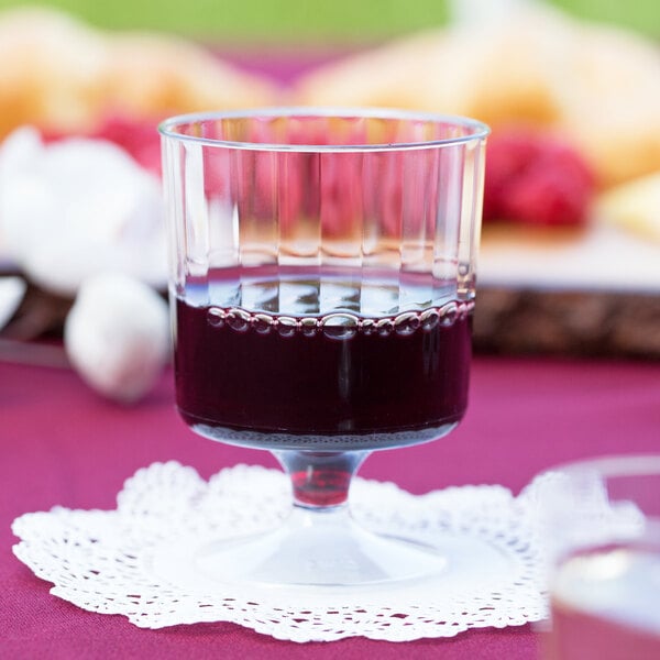 A WNA Comet clear plastic pedestal wine cup filled with red wine on a table with a doily.