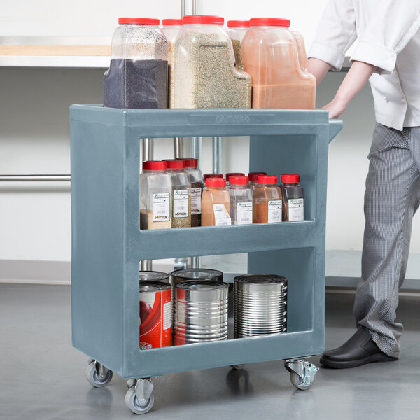 A man pushing a Cambro slate blue plastic service cart with three shelves.