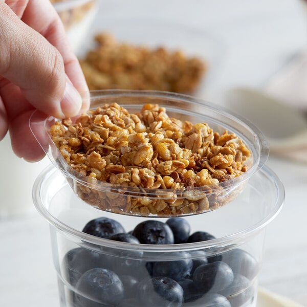 A hand holding a bowl of granola and blueberries over a clear plastic bowl.