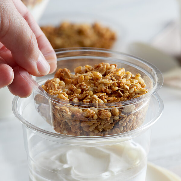 A hand holding a Choice clear plastic parfait insert filled with granola over a white background.