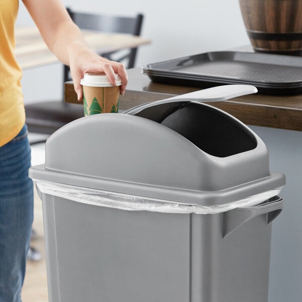 A person throwing a coffee cup into a Lavex gray slim rectangular trash can with a dome lid.