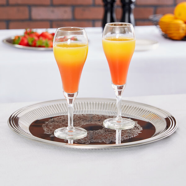 A Vollrath stainless steel round fluted tray holding two glasses of orange and yellow liquid.