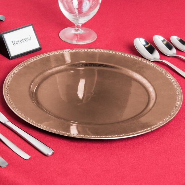 A table setting with a copper charger plate, silverware, and a wine glass on a red tablecloth.